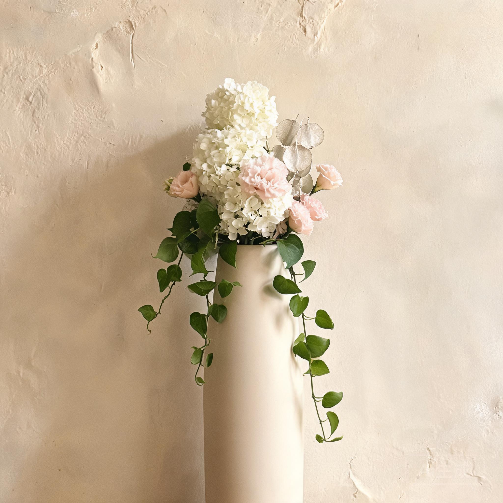 A tall slender ceramic vessel holding a vertical arrangement of white hydrangeas, blush lisianthus, and dried lunaria, photographed against a textured plaster wall.